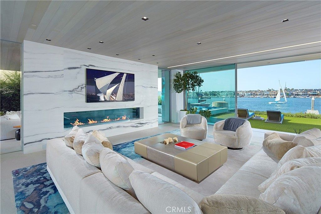 This expansive, modern living room features a large, light-colored sectional sofa and a central coffee table, all arranged to face a striking stone-clad wall with a wide, linear fireplace and a mounted television. The room is defined by its seamless transition to the outdoors through expansive glass walls that offer a panoramic view of the waterfront. The minimalist aesthetic is complemented by a light wood-paneled ceiling and soft, neutral furnishings, creating a serene and luxurious atmosphere.