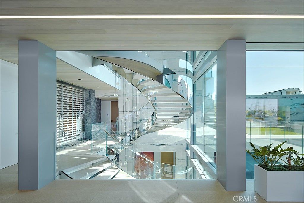 This striking interior space features a dramatic, sculptural spiral staircase encased in glass, serving as the central architectural focal point. The design utilizes clean lines, light-toned wood ceilings, and expansive glass walls that provide a seamless transition between the interior and the outdoor environment. The perspective looks through the glass partition, highlighting the open, airy atmosphere and the sophisticated, modern aesthetic of the home.