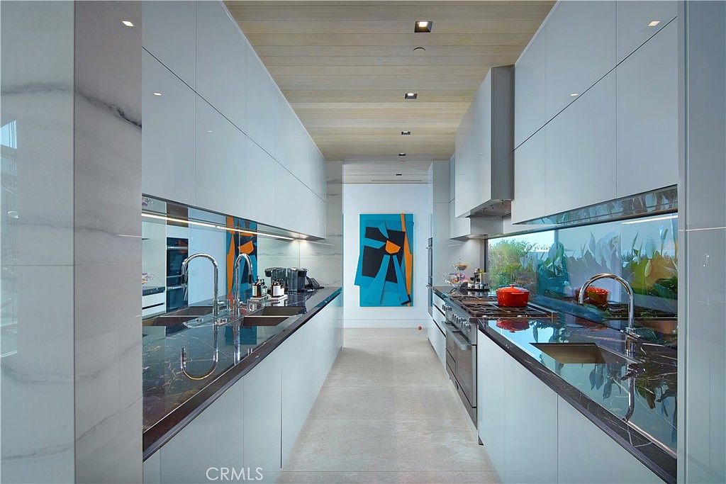 This sleek, galley-style kitchen features high-gloss white cabinetry and dark, patterned stone countertops that create a striking contrast. The space is illuminated by recessed lighting set into a light wood-paneled ceiling, while a vibrant abstract painting adds a pop of color at the far end of the corridor. The perspective looks directly down the center of the kitchen, highlighting the symmetrical layout and the reflective surfaces of the cabinetry and backsplash.