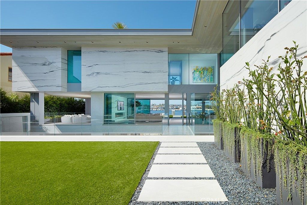 This modern luxury residence features a striking facade clad in large, light-toned stone panels with prominent veining. The home showcases expansive glass walls that provide a seamless transition between the interior living spaces and the outdoor waterfront area. A manicured lawn and a stone paver walkway lead toward the house, framed by sleek planters filled with tall, slender greenery.