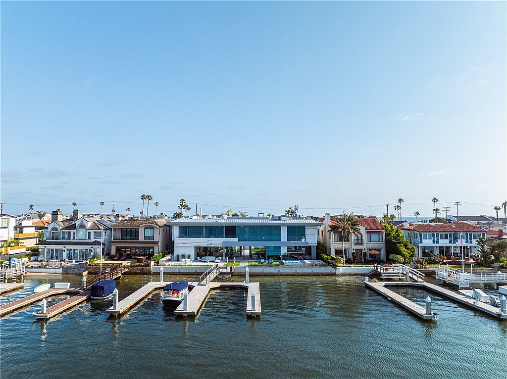 This aerial perspective captures a modern, multi-level waterfront residence situated along a canal, flanked by more traditional homes. The property features a sleek, contemporary design with extensive glass walls and a private dock extending into the water. The scene is set under a clear blue sky, emphasizing the prime location and the seamless integration between the home's outdoor living spaces and the waterway.