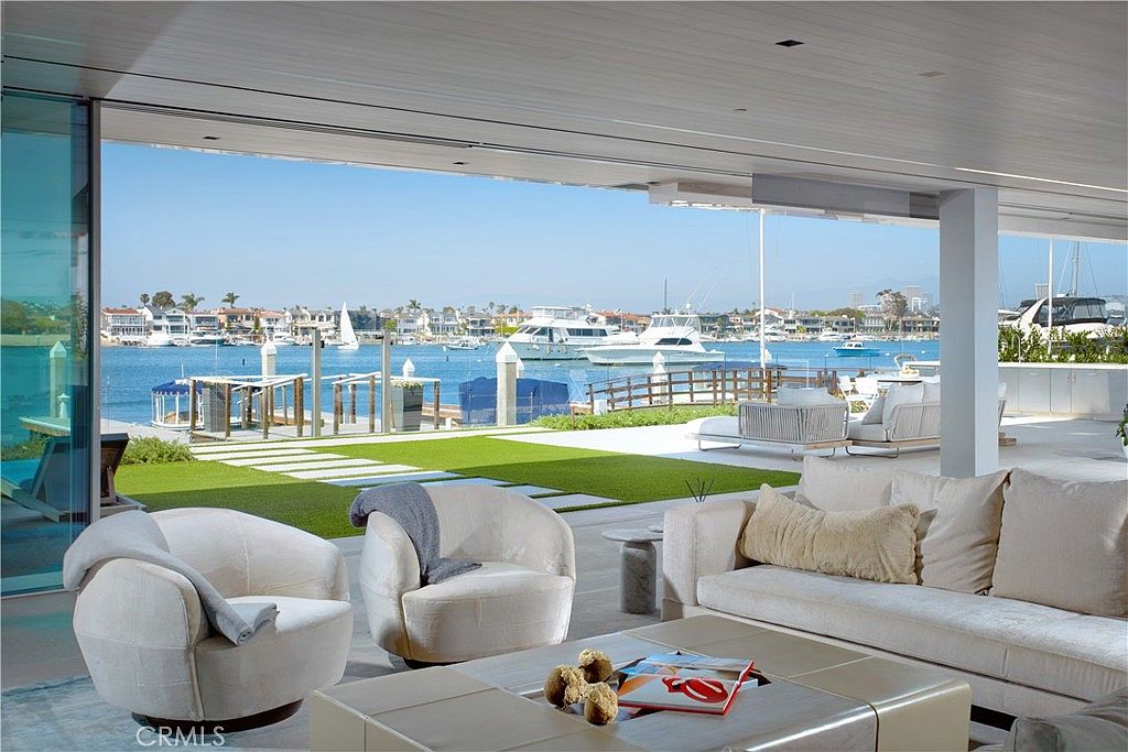 This luxurious indoor-outdoor living space features a seamless transition to a waterfront patio through expansive, retractable glass walls. The interior is furnished with plush, light-toned seating, including two rounded armchairs and a large sofa, centered around a low-profile coffee table. The view beyond the patio highlights a manicured lawn, a private dock, and a serene harbor filled with boats, creating an atmosphere of high-end coastal living.