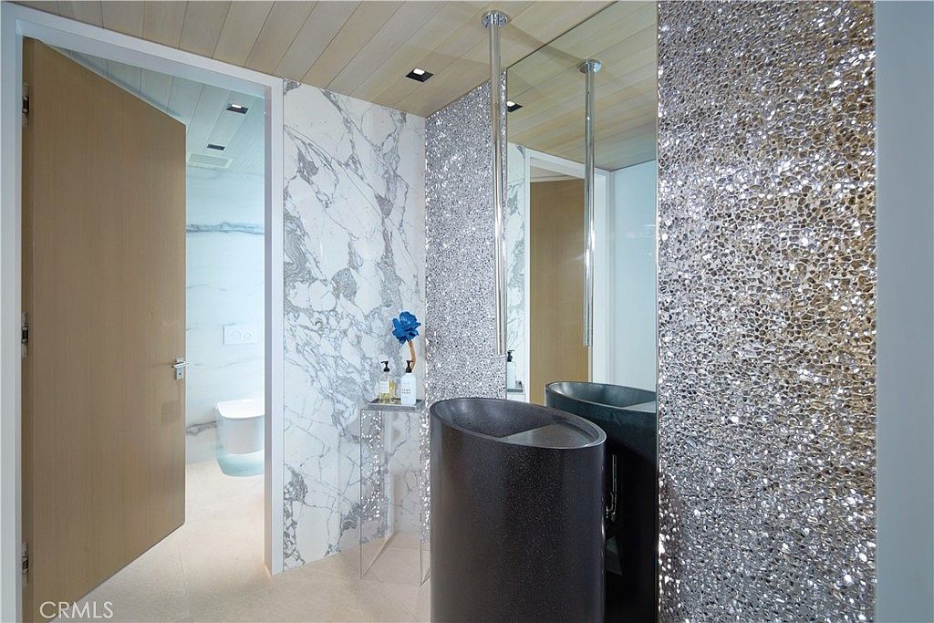 This modern powder room features a striking, dark, cylindrical pedestal sink set against a wall covered in shimmering, textured silver tiles. To the left, a wall of white stone with grey veining provides a clean contrast, while a doorway leads to a secondary bathroom area with a toilet. The ceiling is finished with light-toned wood planks, and a tall, slender mirror reflects the vanity area, creating a sophisticated and high-glamour aesthetic.