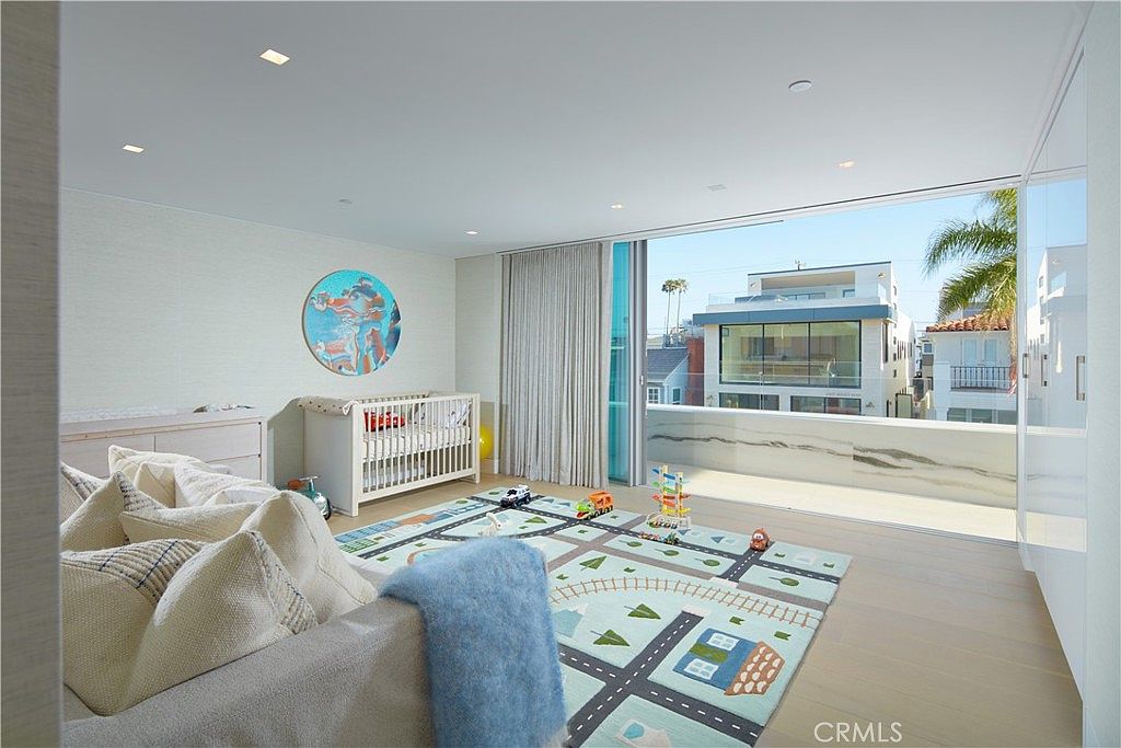 This bright and airy nursery features light wood flooring and a large area rug with a playful road map pattern. A white crib sits against the wall, complemented by a comfortable sofa in the foreground and a circular piece of wall art. The room opens onto a spacious balcony through large sliding glass doors, providing an expansive view of the neighboring coastal-style homes.
