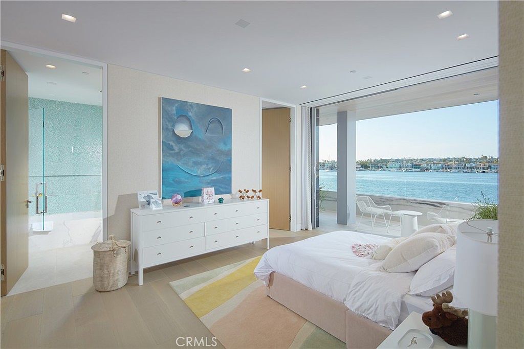 This bright and airy primary bedroom features a contemporary design with light wood flooring and a soft, neutral color palette. A large, open-concept layout connects the sleeping area to an en-suite bathroom with blue mosaic tile, while floor-to-ceiling glass doors open onto a private balcony overlooking the water. The room is accented by a large abstract painting above a white dresser and a plush bed with layered bedding.