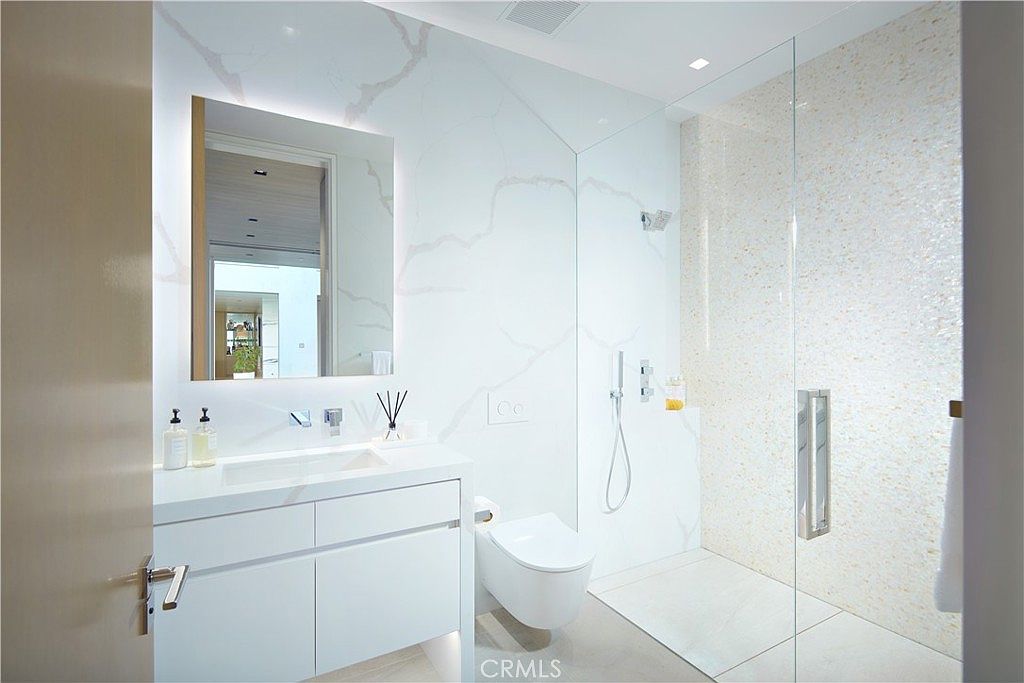 This modern bathroom features a sleek vanity with a white countertop and a large, frameless mirror set against a wall with large-format, light-colored stone tiles. To the right, a glass-enclosed walk-in shower showcases a textured, mosaic-style tile wall, while a wall-mounted toilet is positioned between the vanity and the shower. The space is bright and minimalist, utilizing clean lines and a neutral color palette to create a contemporary aesthetic.