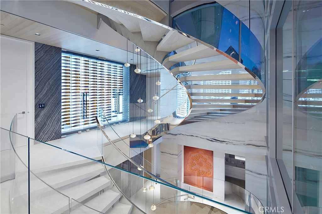 This dramatic interior space features a sweeping, curved staircase with light wood treads and a glass railing that creates an open, airy aesthetic. A striking, multi-tiered pendant light fixture hangs through the center of the staircase, while a large, illuminated wine display wall serves as a sophisticated focal point in the background. The use of reflective surfaces, including glass and polished stone, enhances the luxurious and modern architectural design of the home.