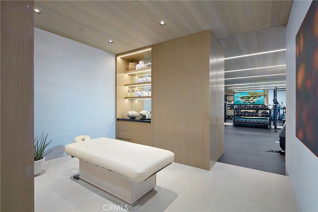 This image showcases a serene massage or wellness room that transitions into a professional-grade home gym. The space features a central massage table, built-in light wood shelving with towels and spa accessories, and a clean, minimalist aesthetic. Through the open doorway, a fully equipped gym with weight racks and a large wall-mounted television is visible, highlighting a comprehensive fitness and relaxation suite.
