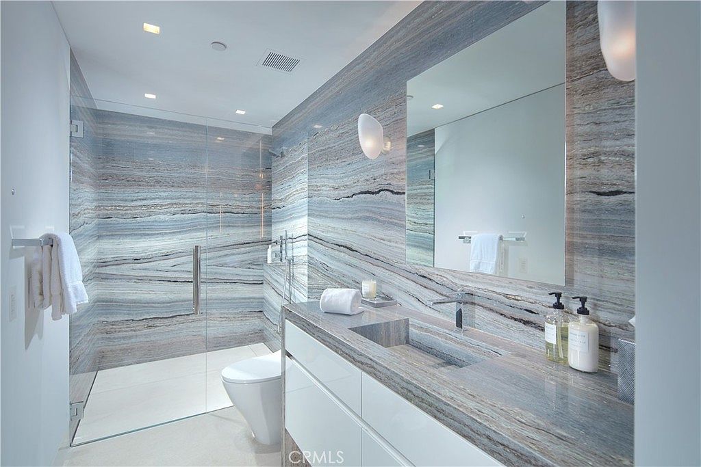 This modern bathroom features striking, heavily veined stone wall cladding that extends throughout the space, creating a cohesive and luxurious aesthetic. The vanity includes an integrated stone sink and a large, frameless mirror, while a glass-enclosed walk-in shower occupies the corner. The perspective is from the doorway, showcasing the sleek, minimalist design and high-end finishes of the room.