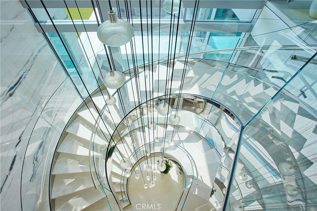 This high-angle, cinematic shot captures a dramatic spiral staircase featuring glass railings and light-colored stone steps. A cluster of spherical pendant lights hangs through the center of the staircase, creating a striking vertical focal point. The perspective emphasizes the architectural geometry and the open, airy feel of the modern interior.