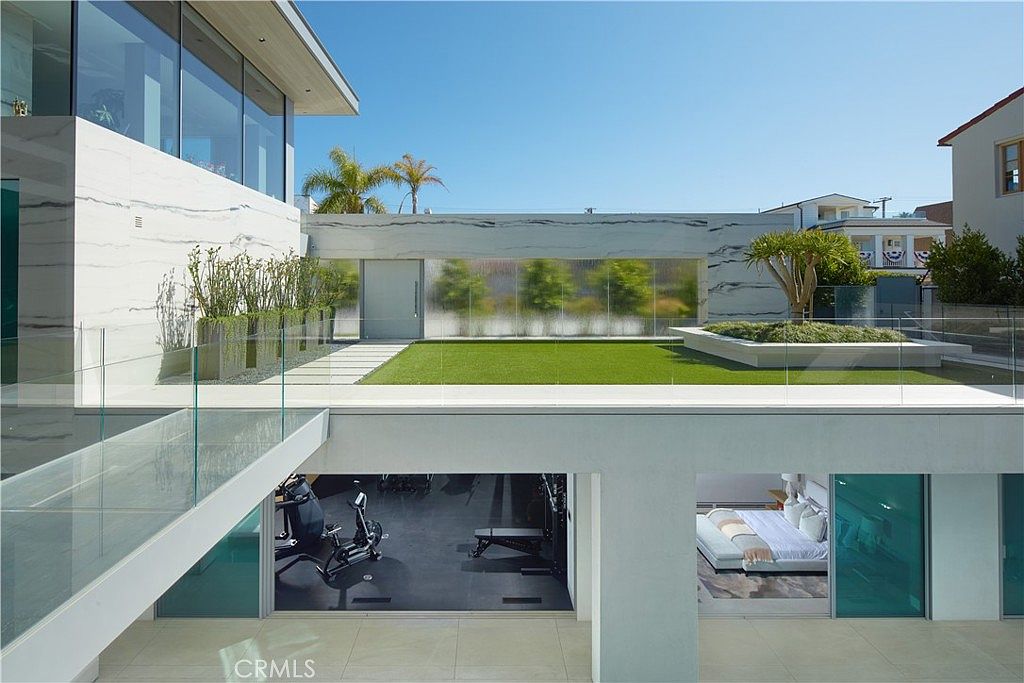 This modern architectural residence features a multi-level design with a prominent upper-level terrace covered in artificial turf and a stone-clad planter. The lower level opens directly to a patio area, revealing a home gym on the left and a bedroom suite on the right through large glass sliding doors. The clean lines, glass railings, and minimalist aesthetic create a sophisticated, high-end contemporary atmosphere.