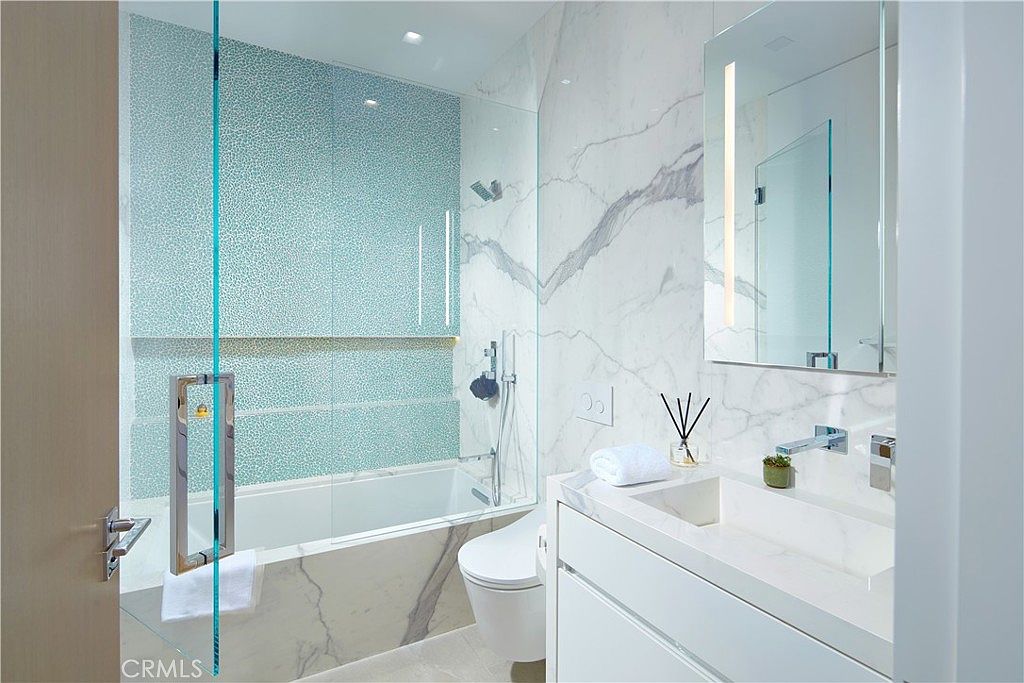 This modern bathroom features a sleek, integrated vanity with a minimalist faucet and a wall-mounted mirror. A glass-enclosed shower and tub combination is highlighted by a striking blue patterned tile accent wall, contrasted against large-format stone wall panels. The space is bright and contemporary, utilizing clean lines and a neutral color palette to create a sophisticated, spa-like atmosphere.