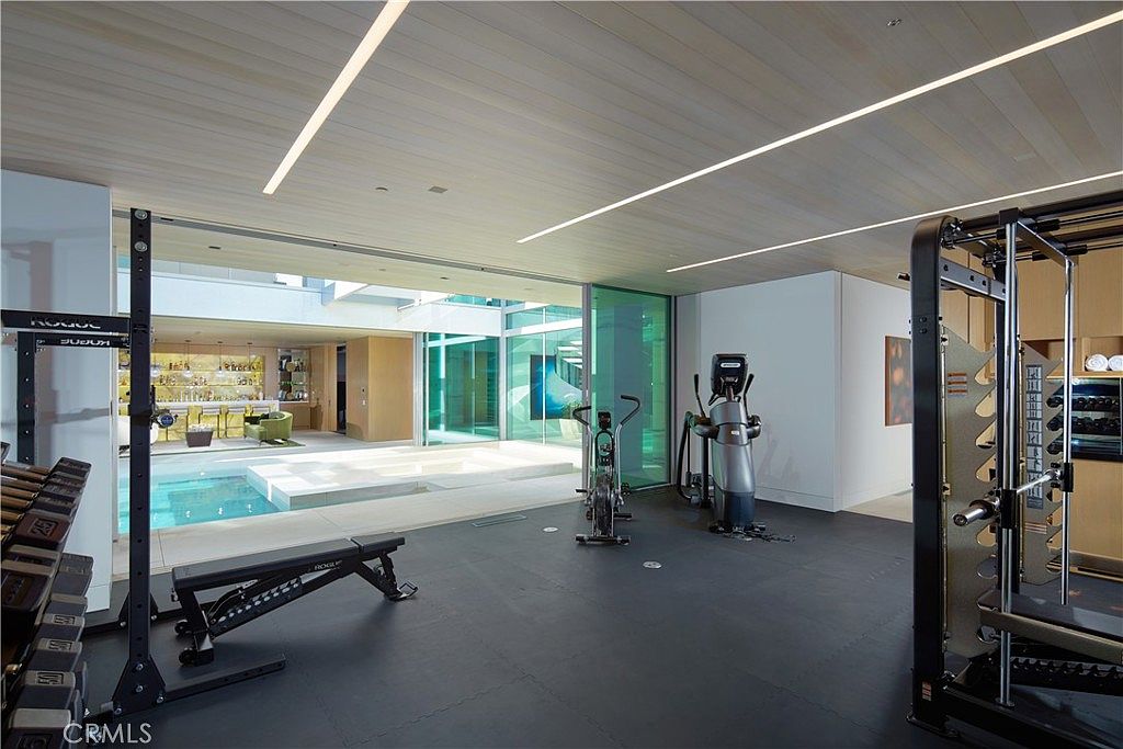 This modern home gym features a professional-grade weight rack, a bench, and cardio equipment set on a dark, durable rubberized floor. The space is defined by its seamless transition to an outdoor pool area through large, sliding glass doors that provide an expansive view of the water and a lounge space beyond. The ceiling is finished with light wood planks and integrated linear lighting, creating a sleek, high-end atmosphere.