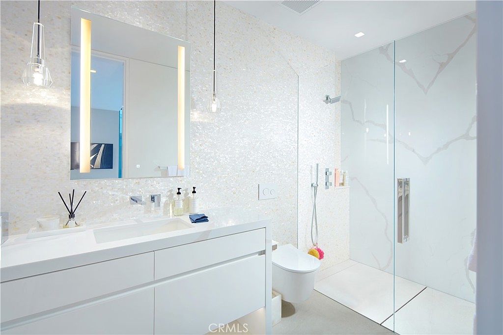 This modern bathroom features a sleek, wall-mounted white vanity with a rectangular sink and a large, illuminated mirror. To the right, a spacious walk-in shower is enclosed by a glass panel and finished with large-format, light-colored stone tiles. The wall behind the vanity is covered in a textured, shimmering mosaic tile, creating a bright and sophisticated atmosphere.