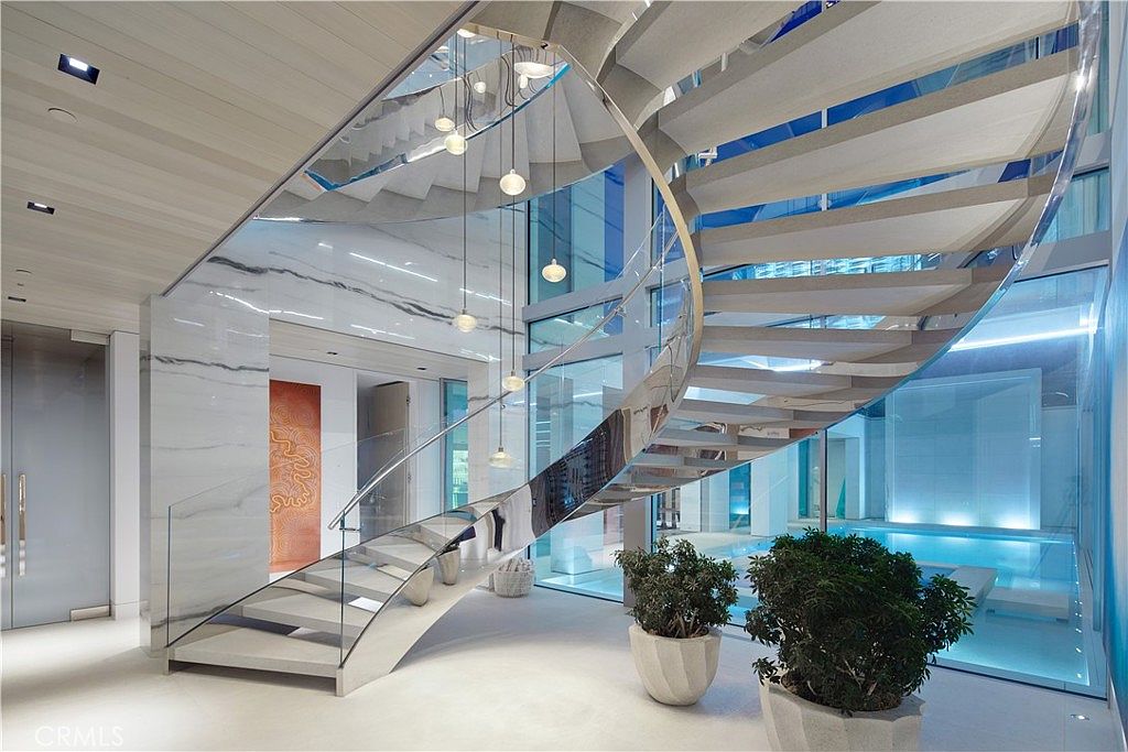 This grand entryway features a striking, curved staircase with a polished metal railing and light-toned treads that serves as the focal point of the space. The area is defined by expansive glass walls that provide a clear view into an indoor pool, creating a seamless blend between the architectural structure and the water feature. A cluster of modern, spherical pendant lights hangs from the ceiling, while large potted plants add a touch of greenery to the minimalist, light-filled interior.