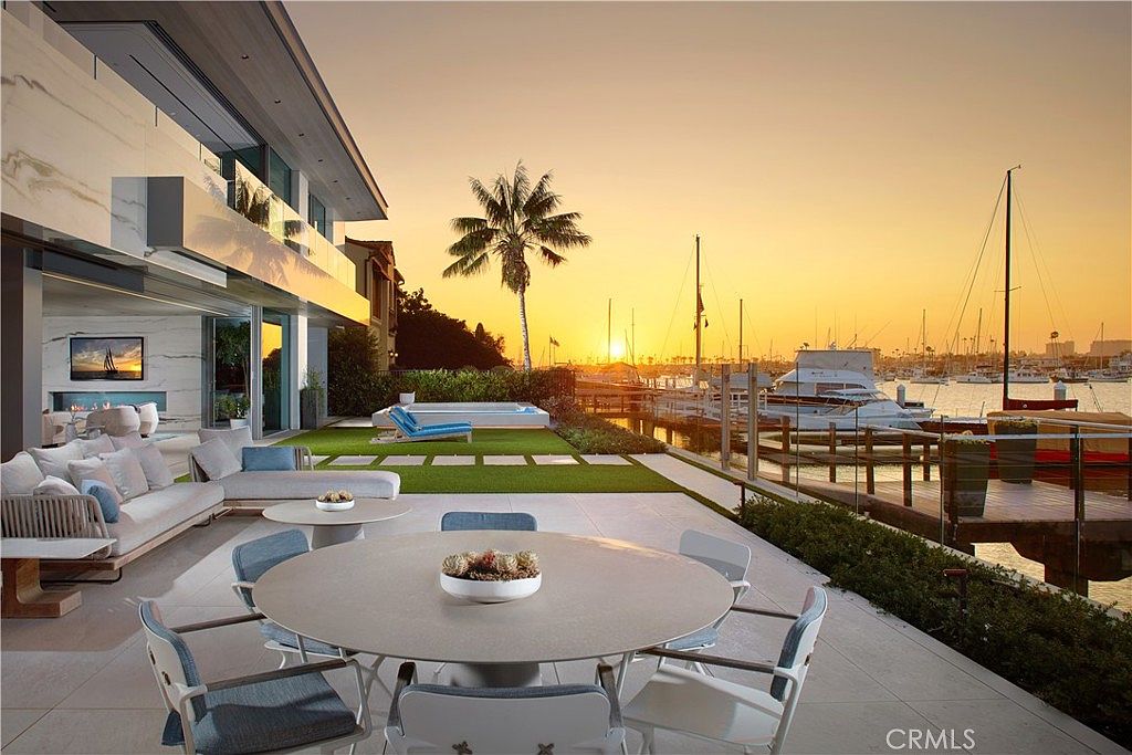 This luxurious waterfront patio offers a seamless transition between indoor and outdoor living, featuring a spacious dining area and comfortable lounge seating. The space overlooks a private dock and a marina filled with boats, all set against a vibrant sunset backdrop. A manicured lawn with a built-in spa sits between the patio and the water, creating an elegant and serene atmosphere.