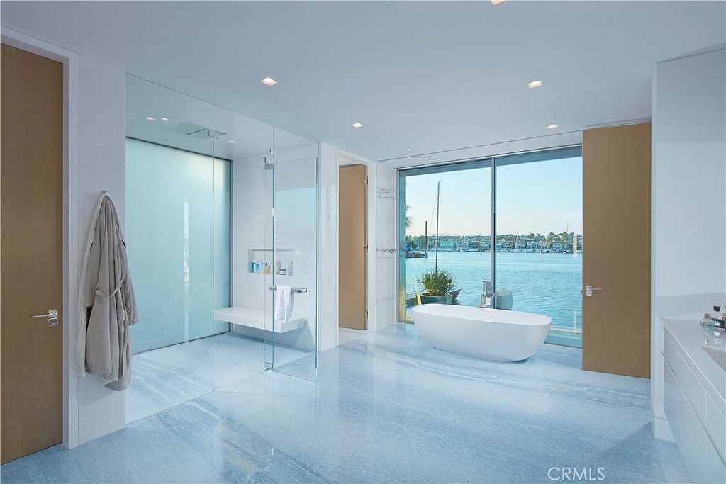 This luxurious primary bathroom features a striking blue-toned stone floor and a freestanding soaking tub positioned in front of large, floor-to-ceiling windows that offer a panoramic view of the harbor. To the left, a spacious walk-in shower is enclosed by glass panels, complemented by a built-in bench and minimalist fixtures. The room is accented with light wood doors and a sleek vanity, creating a serene, high-end atmosphere with a seamless connection to the waterfront scenery.