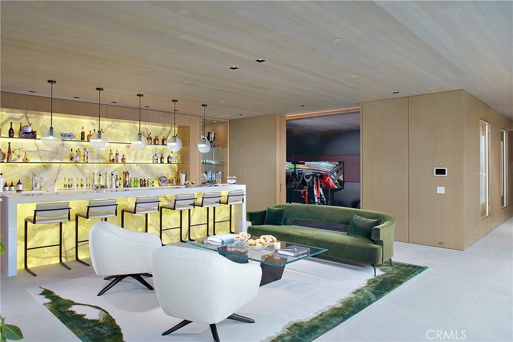 This sophisticated living area features a prominent backlit bar with stone surfaces and multiple pendant lights, creating a warm, glowing focal point. A plush green velvet sofa and two white modern armchairs are arranged around a glass-topped coffee table, set upon a patterned area rug. The room is finished with light wood paneling on the walls and ceiling, offering a sleek, contemporary aesthetic with a view into an adjacent room containing exercise equipment.