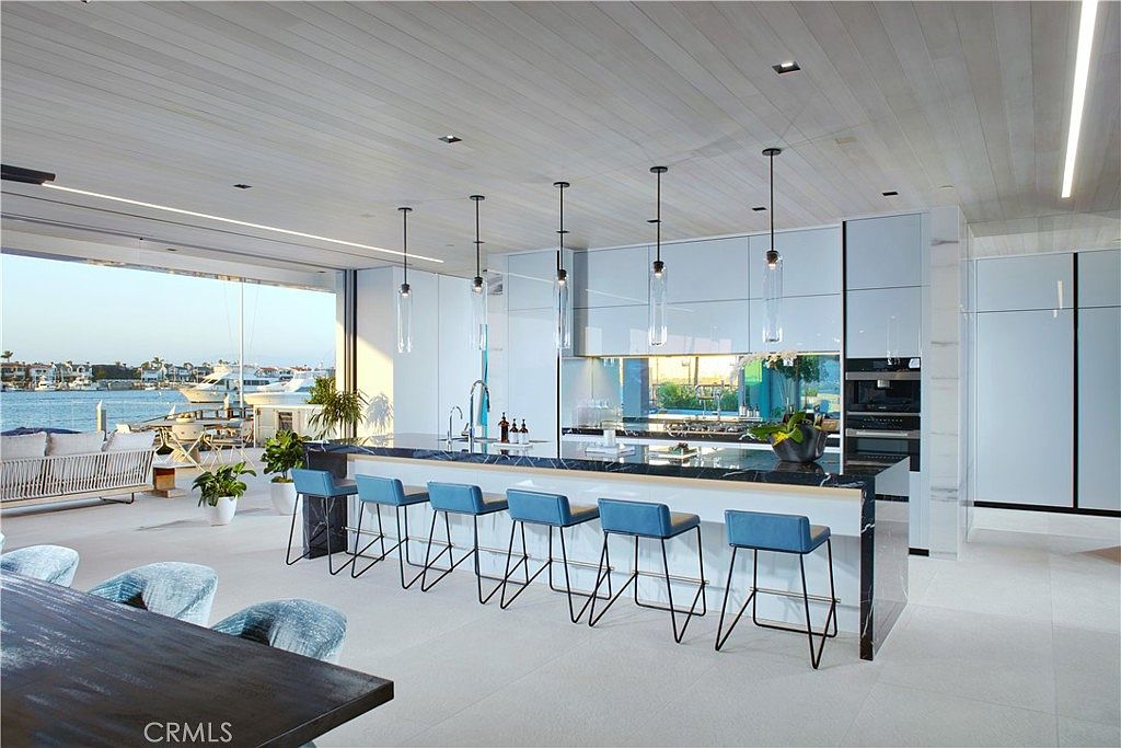 This ultra-modern, open-concept kitchen features a long island with a dark, veined stone countertop and six blue bar stools, seamlessly blending into an expansive outdoor deck area. The space is defined by sleek, high-gloss white cabinetry, minimalist pendant lighting, and a wall of glass that opens directly to a waterfront view with docked boats. The perspective is a wide-angle shot from the dining area, emphasizing the seamless transition between indoor luxury and the coastal environment.