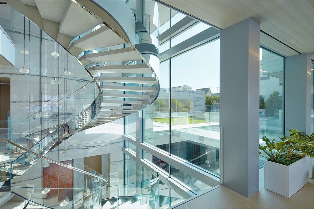 This striking interior space features a dramatic, sculptural spiral staircase with glass railings that serves as the focal point of the room. Large, expansive windows provide an unobstructed view of the outdoor courtyard and pool area, bathing the space in natural light. The minimalist design is characterized by clean lines, neutral stone surfaces, and a sophisticated, modern aesthetic.