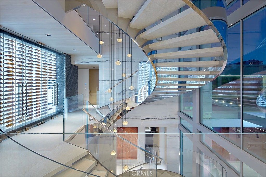 This striking interior space features a dramatic, curved floating staircase with light wood treads and a glass railing that serves as the architectural centerpiece. To the left, a glass-enclosed wine display wall adds a sophisticated, modern touch, while a cluster of delicate pendant lights hangs through the center of the staircase void. The perspective from the landing captures the interplay of light, glass, and clean lines, creating an airy and luxurious atmosphere.