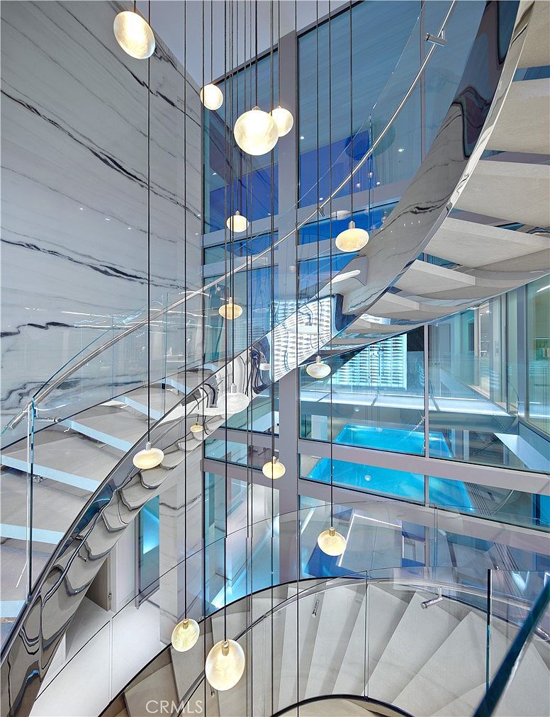 This dramatic, multi-level staircase features a curved design with glass railings and light-colored treads, creating a sense of airy sophistication. A striking cluster of spherical pendant lights hangs vertically through the center of the stairwell, while large glass panels offer a clear view into an adjacent indoor pool area. The space is defined by its clean lines, reflective surfaces, and a high-contrast wall featuring bold, dark veining.