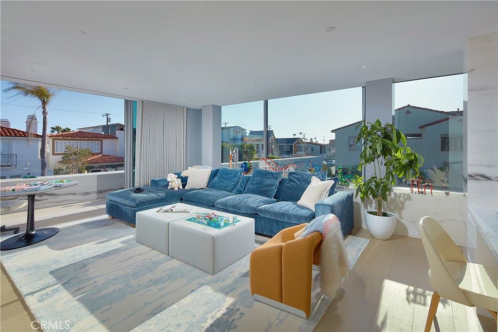 This bright and airy living room features a large blue velvet sectional sofa paired with two light-colored ottomans and a modern orange accent chair. The space is defined by expansive floor-to-ceiling windows that provide panoramic views of the surrounding neighborhood and allow for abundant natural light. A large, light-toned area rug with a subtle blue pattern anchors the seating area, while a potted plant adds a touch of greenery to the minimalist, contemporary interior.