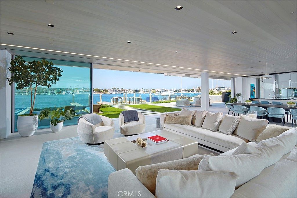 This expansive, open-concept living area features a seamless transition to the outdoors through massive floor-to-ceiling glass walls that reveal a waterfront view. The room is furnished with a large, light-toned sectional sofa, a central square ottoman, and two plush armchairs, all set atop a soft, blue-toned area rug. The minimalist aesthetic is complemented by a light wood-paneled ceiling with recessed lighting and a direct line of sight to a dining area and a private dock on the water.