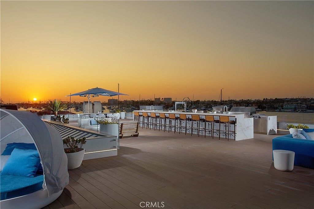 This expansive rooftop deck offers a luxurious outdoor living space with a panoramic sunset view over a harbor filled with boats. The area features a long, stone-topped bar with multiple stools, a modern lounge area with blue seating, and a shaded daybed, all set against a backdrop of a clear, golden-hour sky. The sleek, contemporary design emphasizes an open-air atmosphere perfect for entertaining.