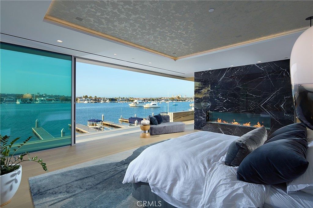 This luxurious primary bedroom features a seamless transition to the outdoors through expansive, wall-to-wall glass panels that reveal a stunning waterfront view. The room is anchored by a bed with crisp white linens and a dark, textured accent wall featuring a sleek, linear fireplace with a full-height stone surround. A plush gray chaise lounge sits near the water's edge, while the ceiling showcases a decorative, metallic-finished recessed design that adds a sophisticated touch to the modern space.