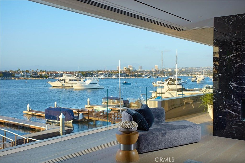 This luxurious outdoor balcony offers a seamless transition from the interior living space to a stunning waterfront view. The area features a modern, plush lounge chair and a decorative wooden side table, all set against a backdrop of a bustling harbor filled with yachts and boats. A glass railing provides an unobstructed perspective of the serene water and the distant city skyline, creating an atmosphere of sophisticated coastal living.
