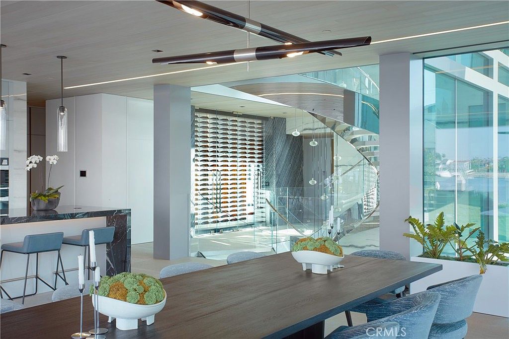 This sophisticated dining area features a long, dark wood table surrounded by upholstered chairs, set against a backdrop of a glass-enclosed wine cellar and a striking spiral staircase. The space is illuminated by a modern, linear light fixture and benefits from natural light streaming through large windows that offer a view of the water. The open-concept design seamlessly connects the dining area to a kitchen island and a contemporary living space, creating an airy and luxurious atmosphere.