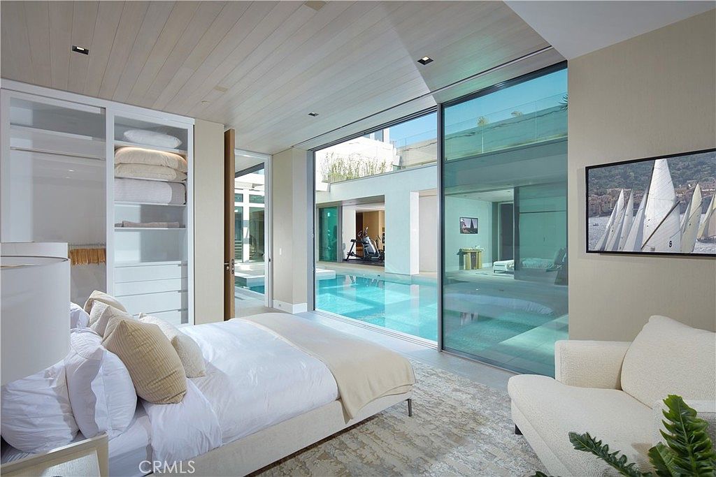 This luxurious primary bedroom features a minimalist design with light wood-paneled ceilings and a neutral color palette. A large glass wall provides a seamless transition to an outdoor pool area, creating a bright and airy atmosphere. The room is furnished with a plush bed, a white armchair, and a built-in closet system, offering a sophisticated and tranquil living space.