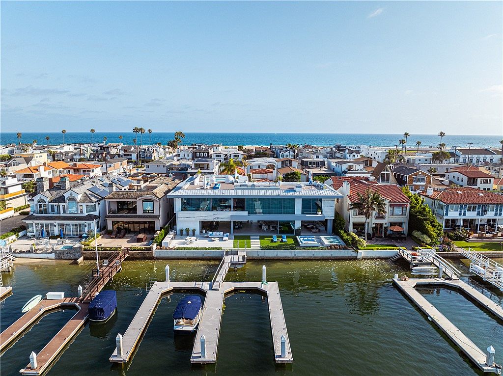 This high-angle aerial view captures a modern, multi-level waterfront residence situated on a canal with private boat docks. The home features a sleek, contemporary design with extensive glass walls, a flat roofline, and a ground-level patio area that transitions into a small lawn and pool. The surrounding neighborhood is densely packed with various architectural styles, all set against the backdrop of a clear blue ocean horizon.