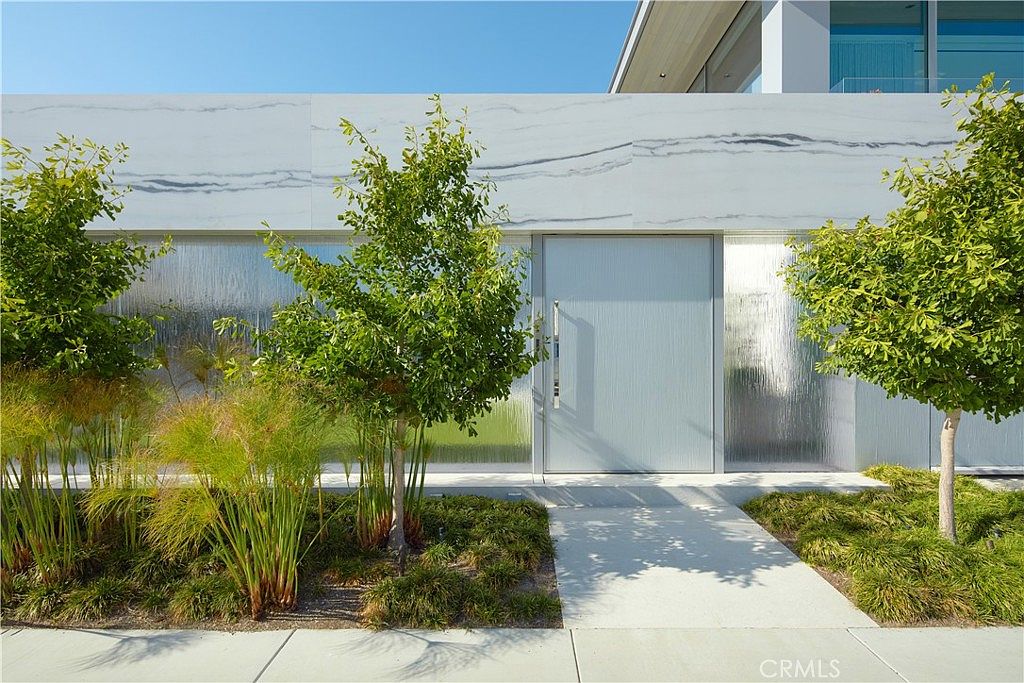 This modern entryway features a minimalist design with a large, light-colored door centered between panels of textured, translucent glass. The facade is composed of smooth, light-toned stone with subtle grey veining, complemented by a manicured landscape of ornamental grasses and small, rounded trees. A clean, concrete walkway leads directly to the entrance, creating a sophisticated and contemporary first impression.