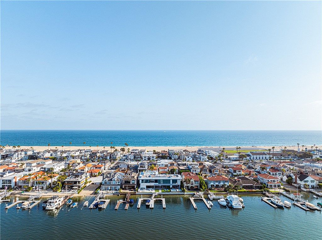 This high-angle aerial perspective captures a luxurious waterfront neighborhood situated between a calm bay and the open ocean. The scene highlights a row of upscale coastal residences, each featuring private boat docks and slips extending into the water. The clear blue sky and expansive horizon emphasize the prime location, showcasing a blend of modern architecture and classic beachside living.