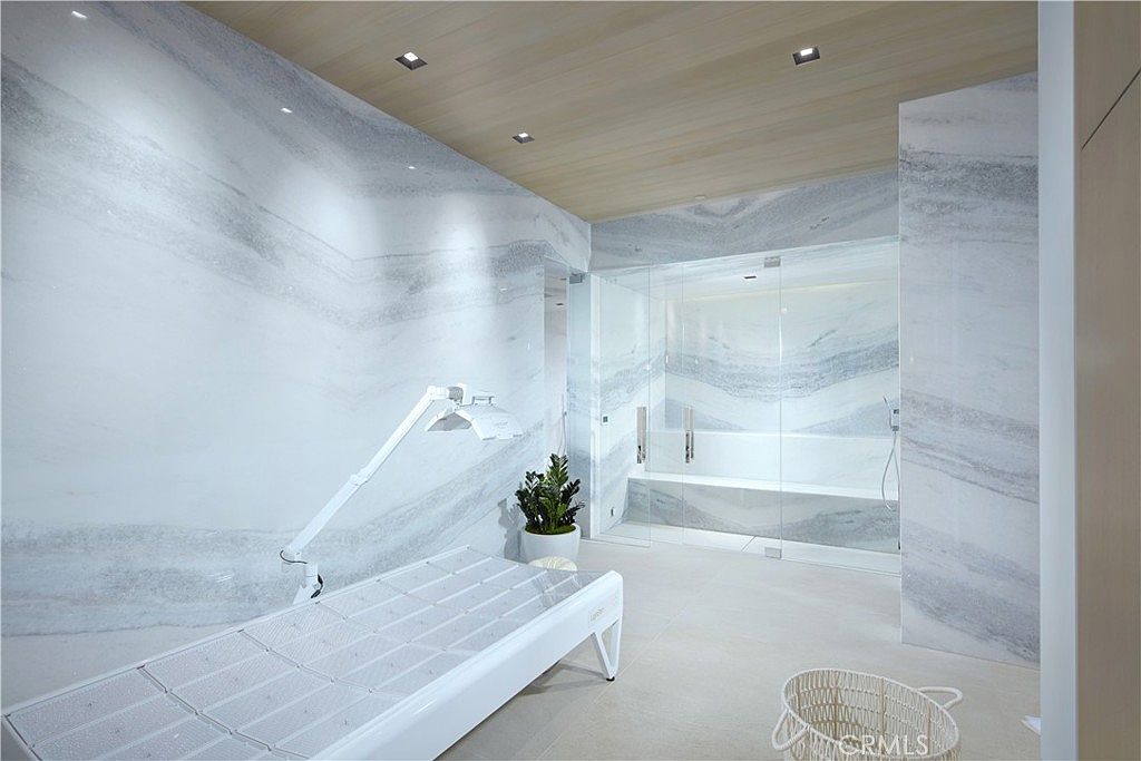 This luxurious wellness room features striking, large-format stone wall cladding with dramatic veining that extends into a glass-enclosed steam shower area. A professional-grade treatment table with an adjustable overhead lamp occupies the center of the space, which is finished with light-toned flooring and a warm wood-paneled ceiling. The room offers a serene, spa-like atmosphere designed for relaxation and personal care.