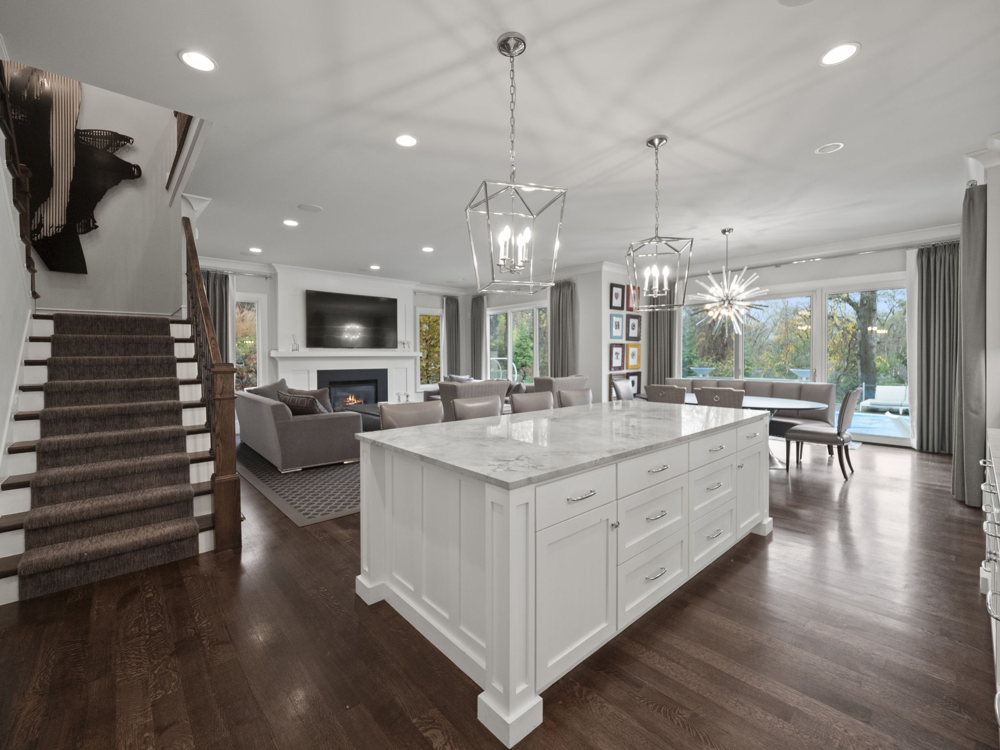 This is a wide-angle shot of a modern, open-concept kitchen and living area. The kitchen features a large white island with a marble countertop, pendant lighting, and dark hardwood floors. The living area includes a fireplace, comfortable seating, and large windows overlooking the backyard, creating a bright and inviting space.