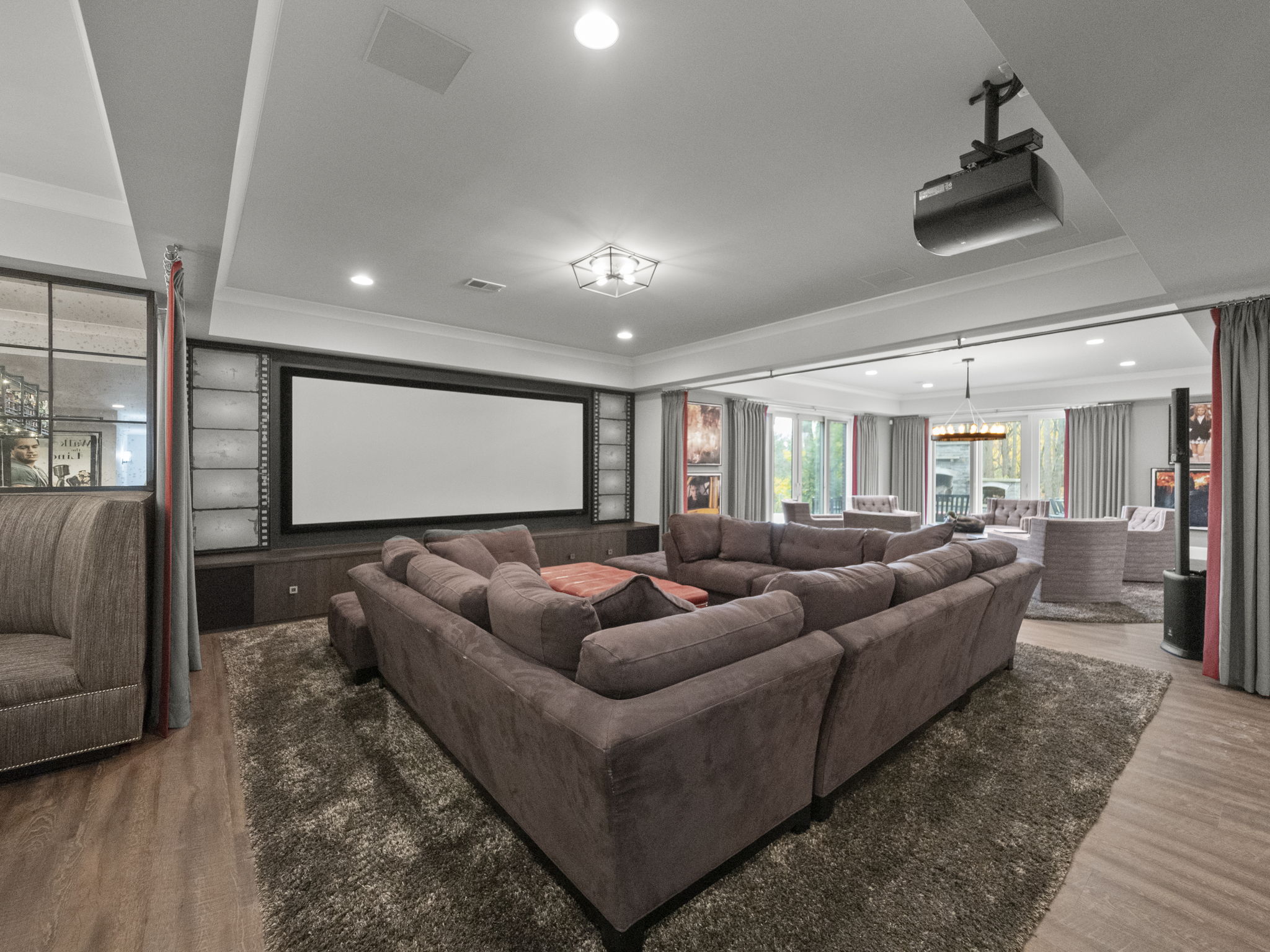 This is a spacious home theater room featuring a large sectional sofa, a projection screen, and a ceiling-mounted projector. The room is carpeted and has a neutral color palette, creating a comfortable and immersive viewing experience. The perspective is from the back of the room, looking towards the screen, capturing the full scale of the space.