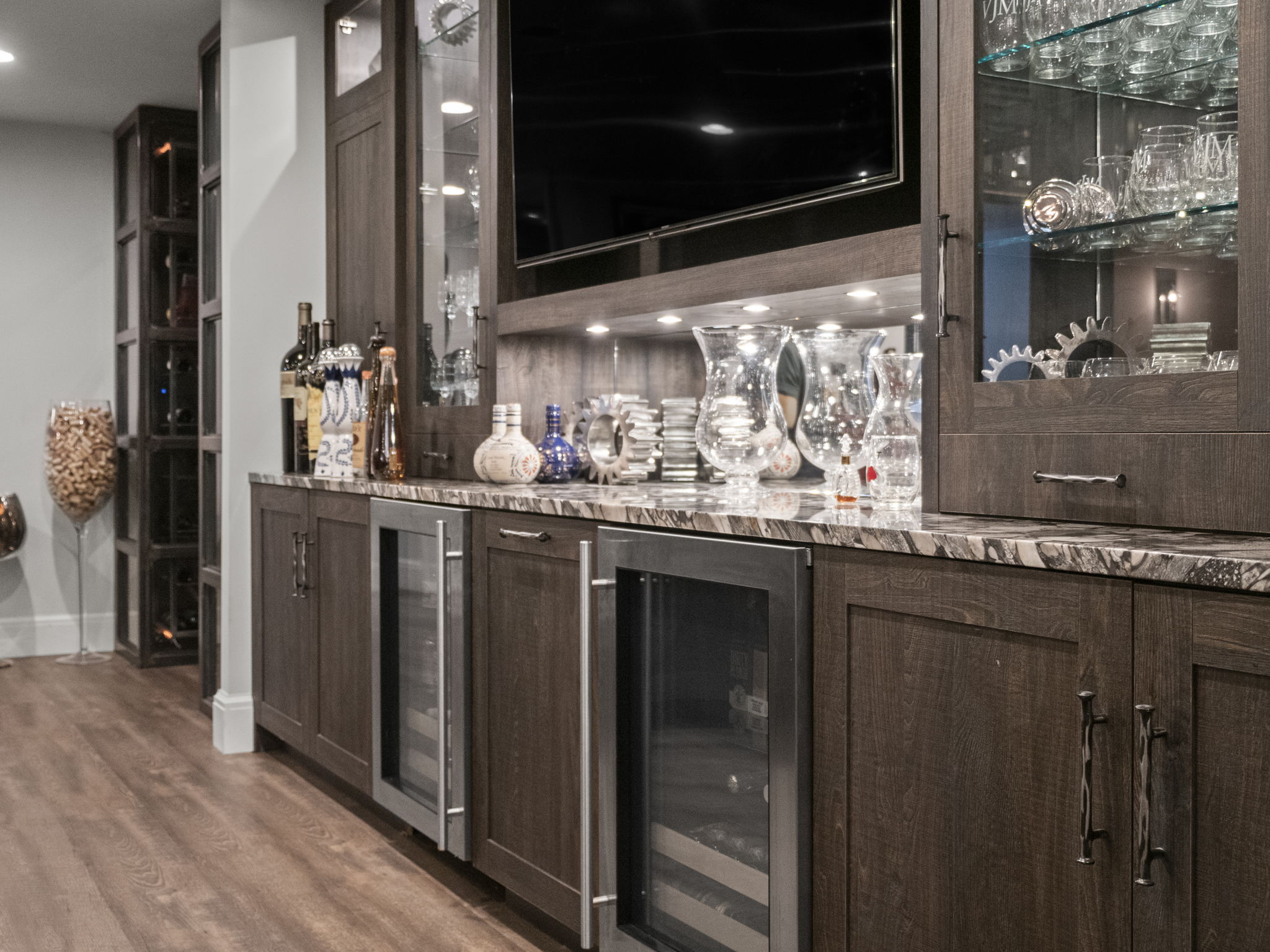 This image showcases a sophisticated wine cellar or bar area, featuring dark wood cabinetry, marble countertops, and glass-front refrigerators for wine storage. The built-in design includes display shelves for glassware and decorative items, illuminated by subtle lighting, creating an elegant and inviting space for entertaining. A large television is mounted above the counter.