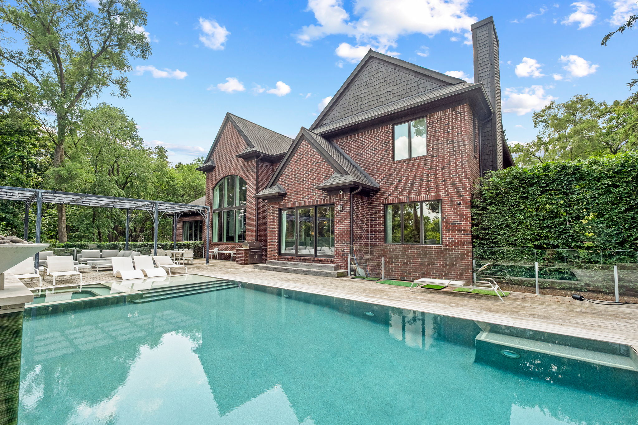 This image showcases a luxurious backyard pool area. The pool features a modern design with integrated steps and a wooden deck surround. Lounge chairs and an outdoor seating area are visible, suggesting a relaxing and upscale outdoor living space, complemented by the brick facade of the house.