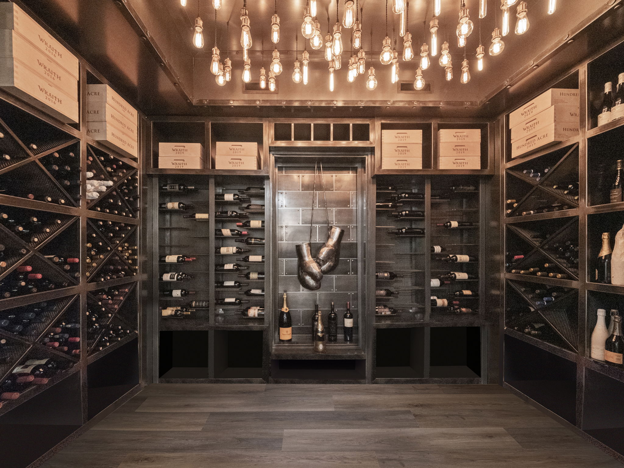 This is a well-stocked wine cellar featuring extensive wine racks filled with bottles. The room is illuminated by numerous vintage-style light bulbs hanging from the ceiling, creating a warm and inviting ambiance. A central display area showcases additional bottles and decorative elements, adding to the cellar's sophisticated appeal.