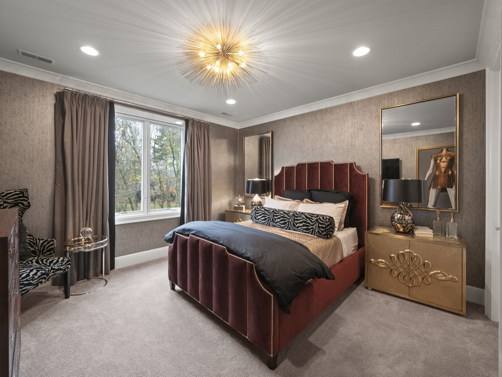 This is a luxurious primary bedroom featuring a red velvet upholstered bed with a black duvet and decorative pillows. The room is decorated with textured wallpaper, a gold accent cabinet, and a statement chandelier. The overall impression is elegant and sophisticated, creating a comfortable and stylish retreat.