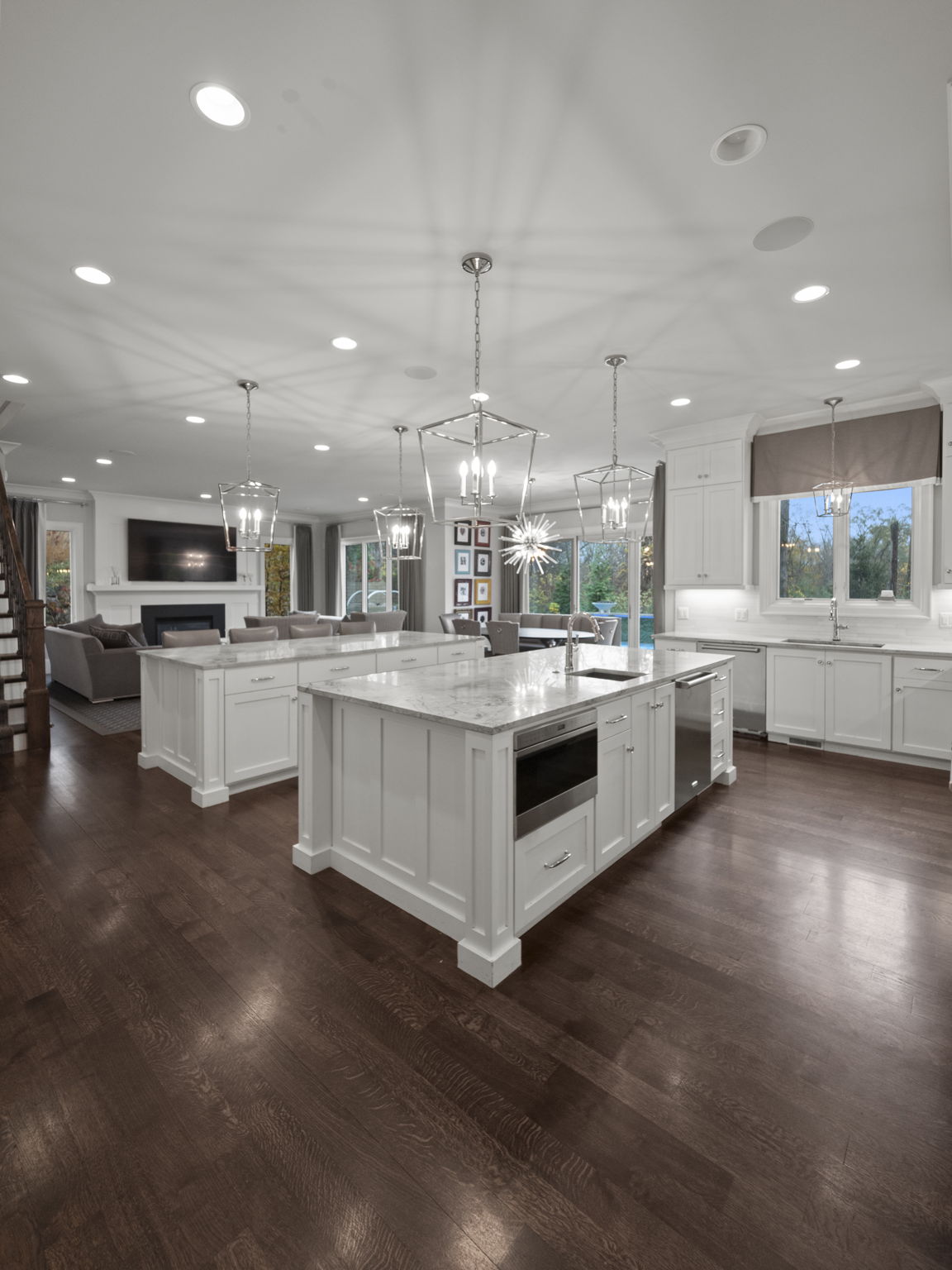 This is a wide-angle shot of a modern, open-concept kitchen that flows into a living area. The kitchen features two large islands with white cabinetry and marble countertops, stainless steel appliances, and pendant lighting. The living area includes a fireplace and comfortable seating, creating a cohesive and inviting space.