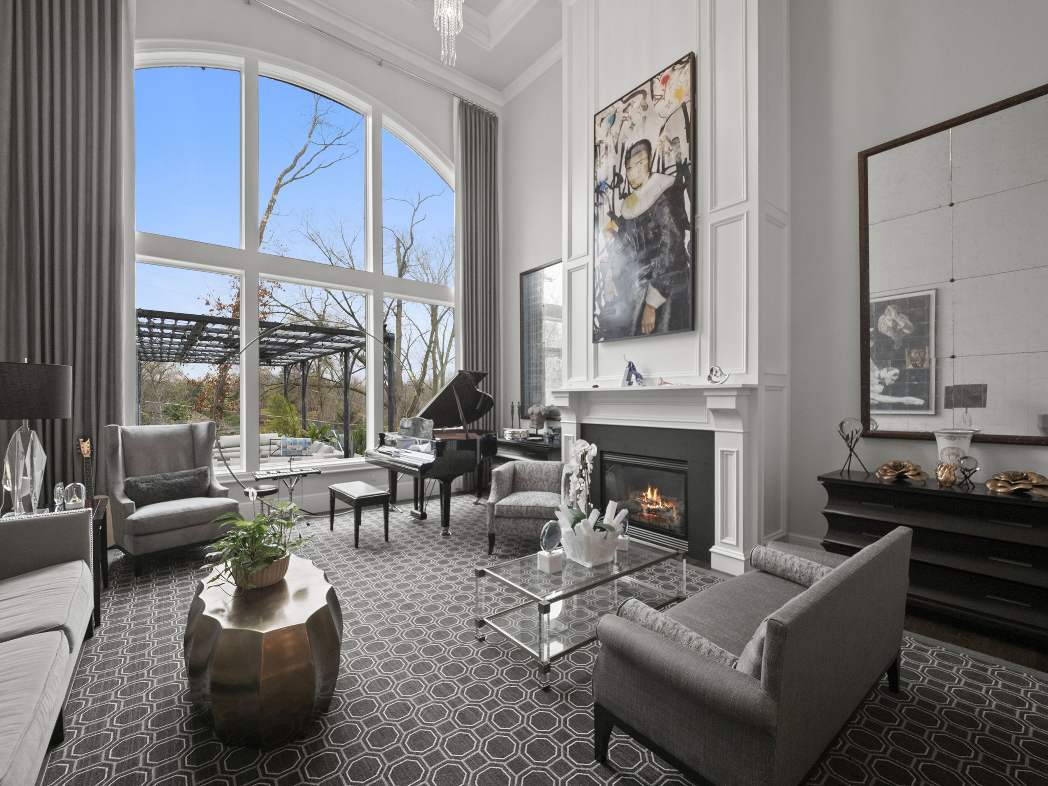 This is an elegant living room featuring a grand piano, a fireplace, and large windows offering natural light. The room is decorated in neutral tones with gray furniture and a patterned rug, creating a sophisticated and inviting atmosphere. Artwork and decorative items add personality to the space.