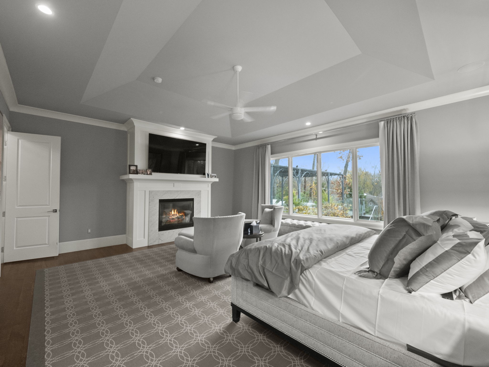 This is a spacious primary bedroom featuring a fireplace with a white mantel and a television above it. The room has a neutral color palette with gray walls and a patterned carpet. Large windows provide natural light and a view of the outdoors, complemented by light gray curtains. The bed is neatly made with white linens and gray pillows, and there are two armchairs near the window, creating a cozy and inviting atmosphere.