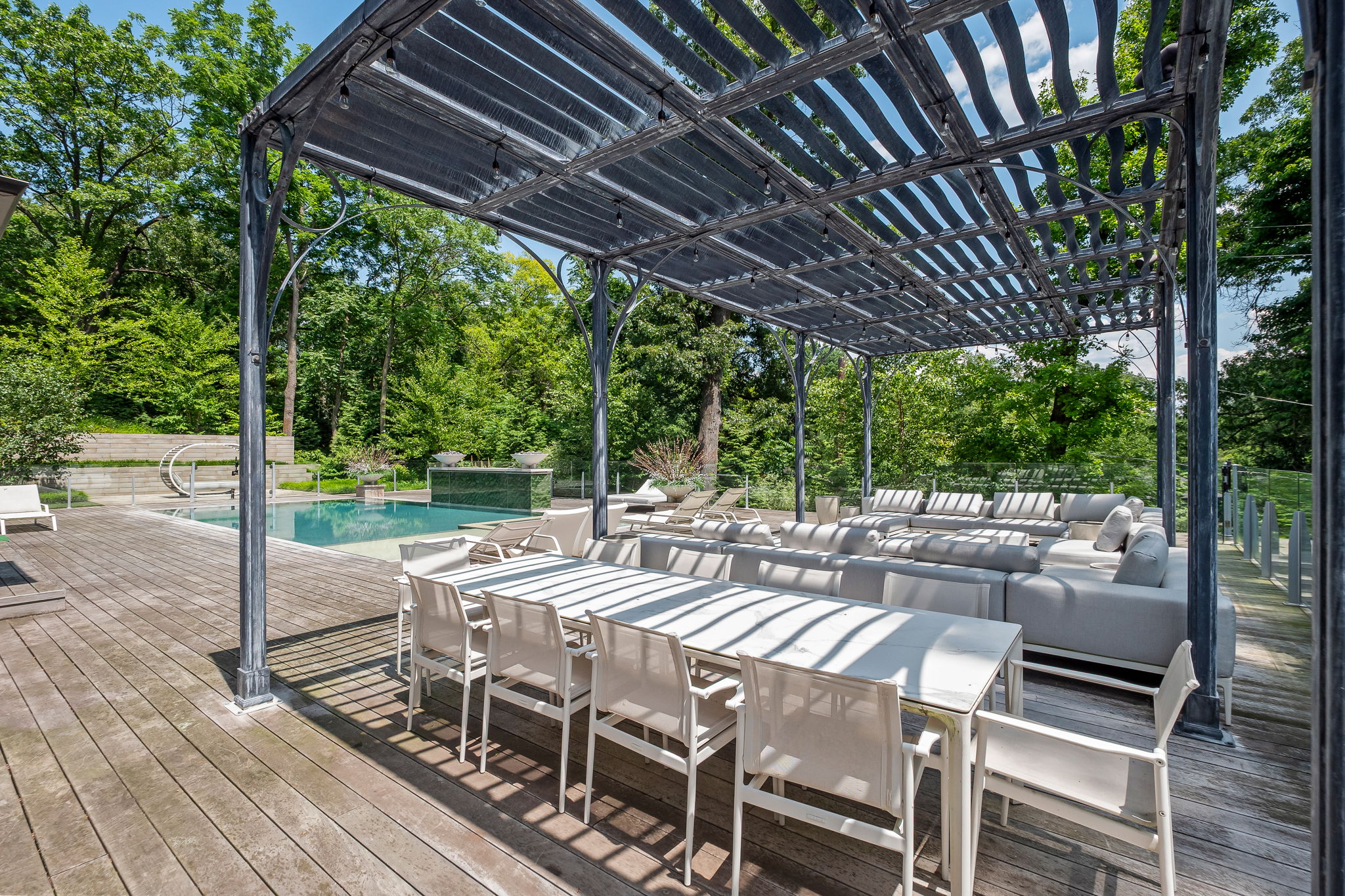 This image showcases a luxurious outdoor living space featuring a spacious wooden deck, a modern pergola, and comfortable lounge seating. A long dining table with chairs is set for entertaining, and a swimming pool is visible in the background, surrounded by lush greenery. The overall impression is one of relaxation and upscale outdoor living.