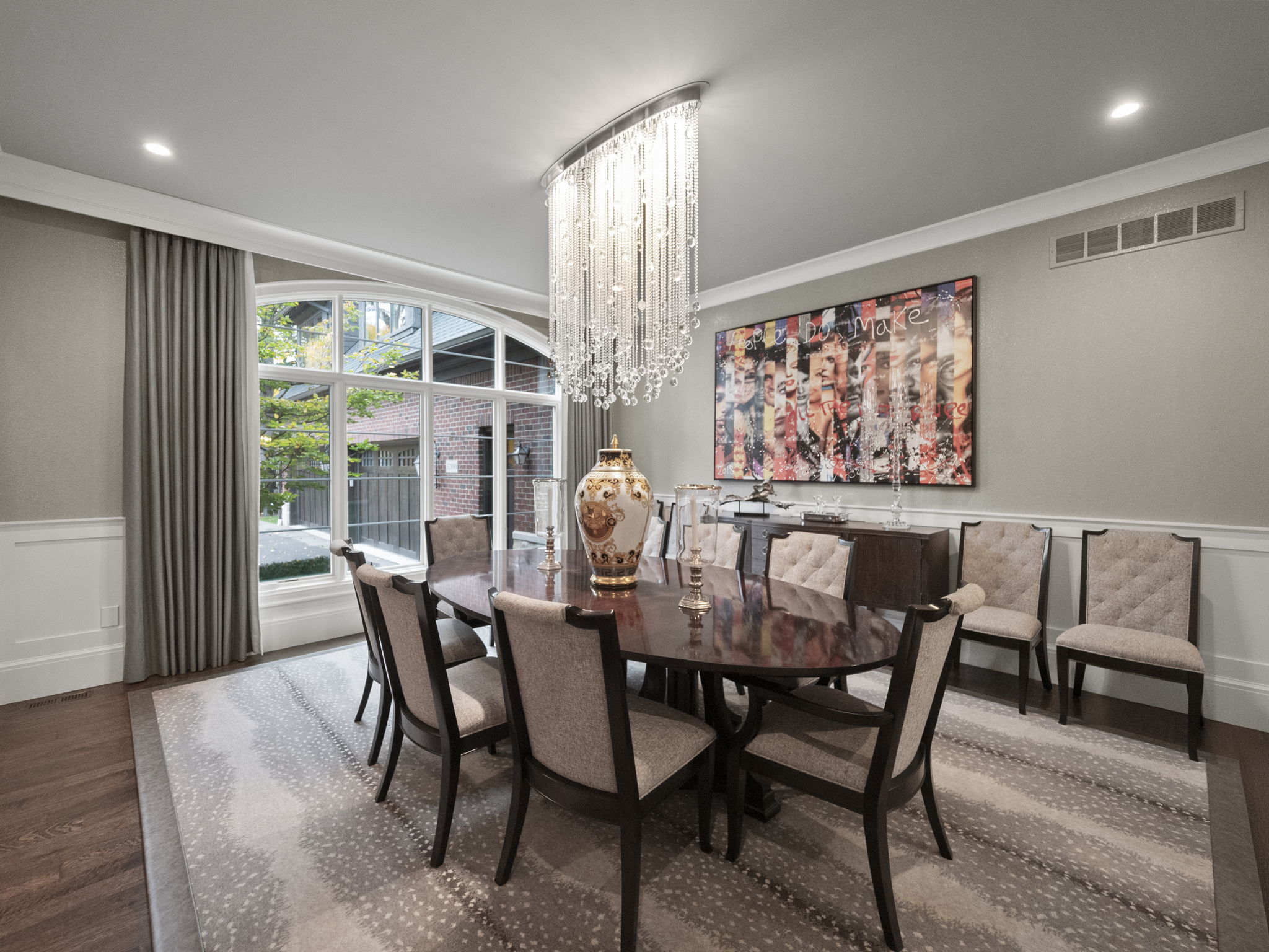 This is an elegant dining room featuring a large oval table with seating for eight, illuminated by a stunning crystal chandelier. A decorative vase sits in the center of the table, and an abstract art piece hangs on the wall. The room is decorated in neutral tones with a patterned rug, creating a sophisticated and inviting atmosphere.