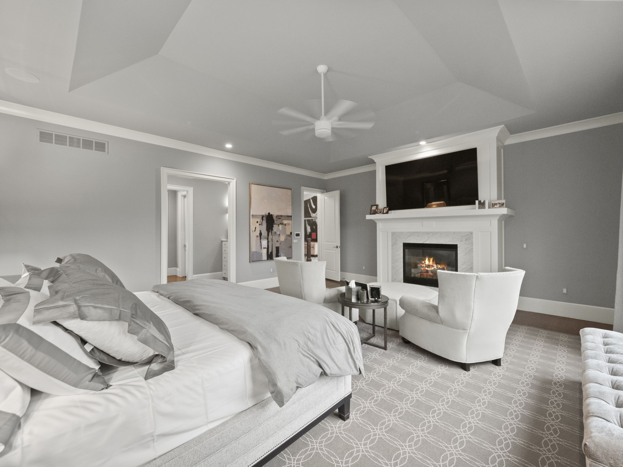 This is a spacious primary bedroom featuring a king-size bed with neutral-toned bedding, a fireplace with a white mantel and a television above it, and two armchairs with a small table between them. The room is decorated in a calming gray color palette with white trim, and the patterned carpet adds a touch of elegance. The perspective is from the foot of the bed, looking towards the fireplace.