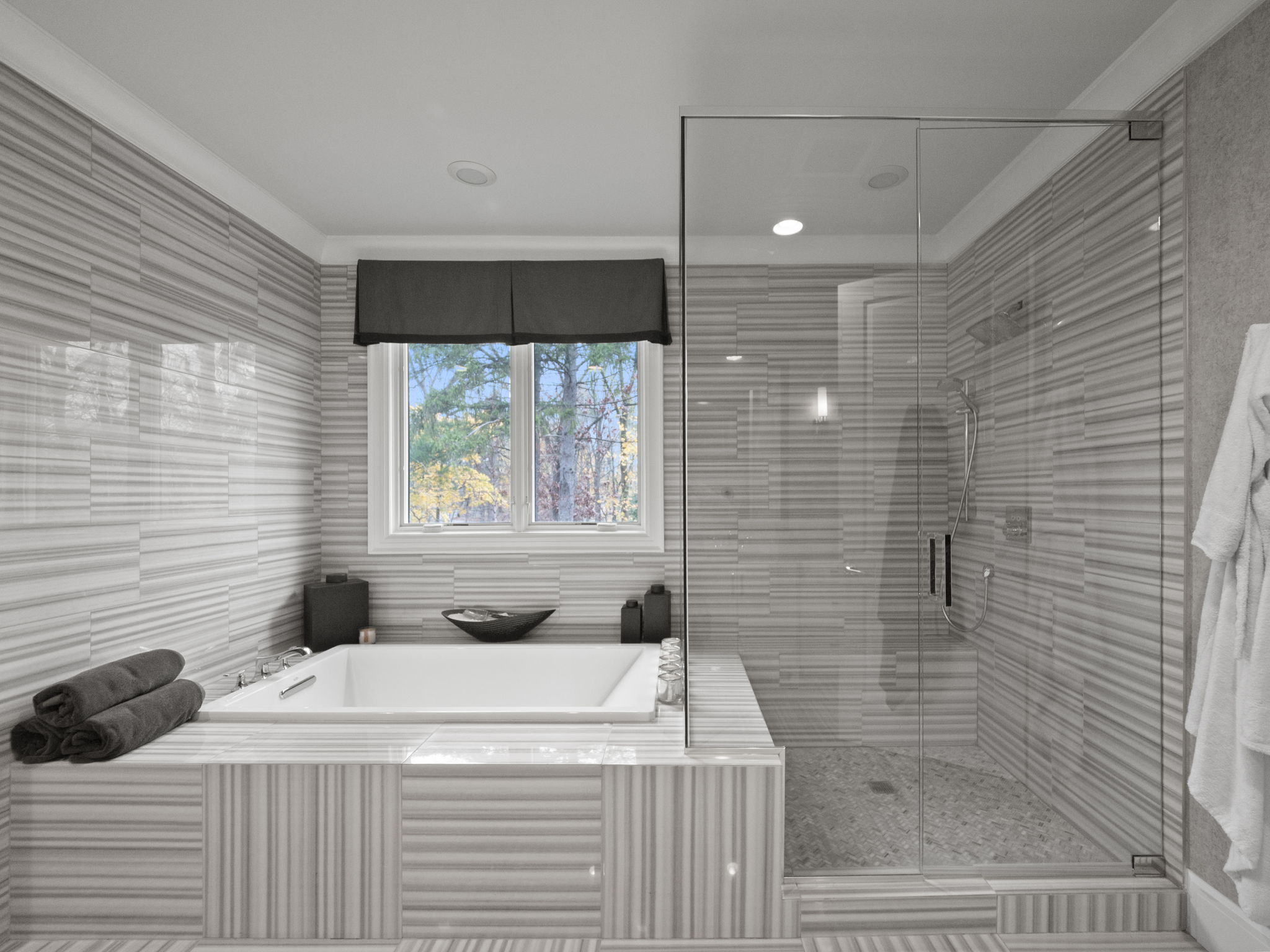 This is a luxurious primary bathroom featuring a soaking tub with a window view and a glass-enclosed shower. The walls and floor are covered in horizontally striped gray and white tiles, creating a modern and clean aesthetic. The perspective is a wide shot, capturing the entire bathroom layout and highlighting the spa-like atmosphere.